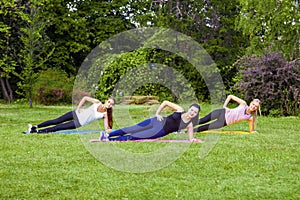 Group of three beautiful healthy slimy woman doing exersices on the green grass in the park, side palnk, looking at camera with