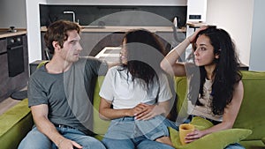Group of three friends one man and two women talking, laughing together while sitting on the couch. Mixed race young