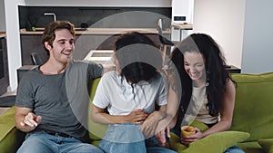 Group of three friends one man and two women talking, laughing together while sitting on the couch. Mixed race young