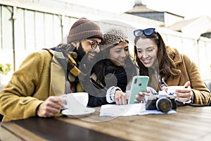 group of three happy friends using phone in outdoor cafe on sunny day - cheerful holidays concept