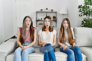 Group of three hispanic girls sitting on the sofa at home afraid and shocked with surprise expression, fear and excited face