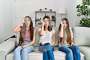 Group of three hispanic girls sitting on the sofa at home doing ok gesture shocked with surprised face, eye looking through