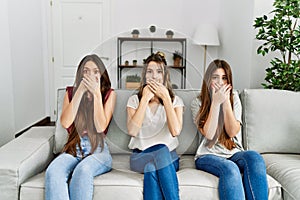 Group of three hispanic girls sitting on the sofa at home shocked covering mouth with hands for mistake