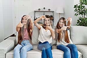 Group of three hispanic girls sitting on the sofa at home smiling making frame with hands and fingers with happy face