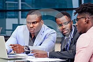 A group of three stylish African American men entrepreneurs in fashion business suits working sitting at table with