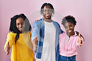 Group of three young black people standing together over pink background smiling friendly offering handshake as greeting and