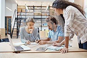 Group of three young multi-ethnic successful business people sitting in coworking space, talking about new project of photo