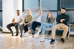 Group of tired five young diverse multiethnic men and women waiting job interview in queue line row in modern office