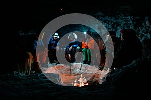 group of tourists in bright ski suits sit around a campfire in winter evening.