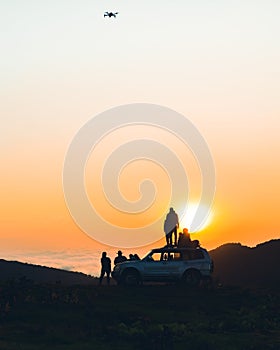 Group of travelers content creators stand by 4wd vehicle together outdoors in nature adventure watch sunset over horizon over