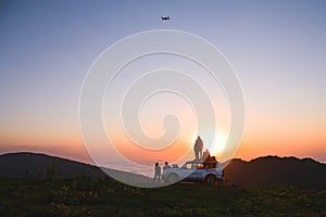 Group of travelers content creators stand by 4wd vehicle together outdoors in nature adventure watch sunset over horizon over