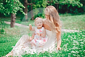 Group of two people, white Caucasian mother and baby girl child in white dress sitting playing in green summer park forest outside