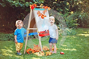 Group of two white Caucasian toddler children kids boy and girl standing outside in summer autumn park by drawing easel