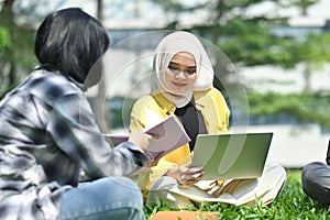 Group of university student discussing their home assignment, using laptop on green grass in campus. Youth lifestyle