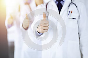 Group of unknown doctors stand as a team with thumbs up in a sunny hospital office. Physicians ready to examine and help