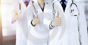 Group of unknown doctors stand as a team with thumbs up in a sunny hospital office. Physicians ready to examine and help