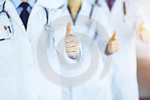 Group of unknown doctors stand as a team with thumbs up in a sunny hospital office. Physicians ready to examine and help