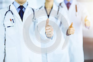 Group of unknown doctors stand as a team with thumbs up in a sunny hospital office. Physicians ready to examine and help