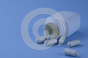 A group of white capsules of medicine spilled out of a white jar on a blue background.