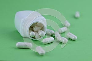 A group of white capsules of medicine spilled out of a white jar on a green background.