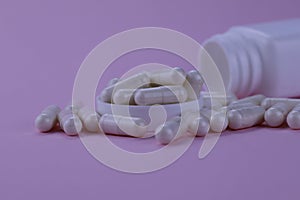 A group of white capsules of medicine spilled out of a white jar on a pink background.