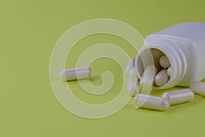 A group of white capsules of medicine spilled out of a white jar on a yellow background.