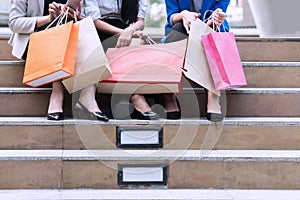 Group woman sitting on stairs and chatting after buying presents. Three girls with high heels and shopping bags in city.