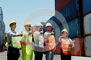 Group of worker team with helmet and safety vest stand in line and give thumbs up at logistic shipping container yard. Woman and