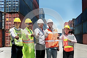 Group of worker team with helmet and safety vest stand in line and give thumbs up at logistic shipping container yard. Woman and