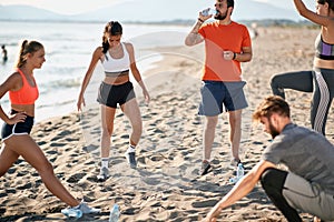 Group of young adults doing exercises on sandy beach. beardy guy drinking water from a plastic bottle