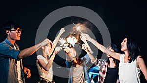 Group of young Asian college student friends lit light sparkler, sing and dance together at beach camping tent