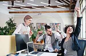 Group of young cheerful businesspeople using laptop in office, expressing excitement.