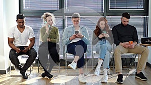 Group of young diverse ethnicity candidates using mobile phones while waiting job interview in modern office lobby.