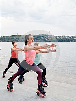 A group of young fit slim women in kangoo jumps, training in front of city lake in summer. Four girls, wearing colorful sports
