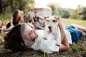 A group of young friends with a dog sitting on grass on a roadtrip through countryside.