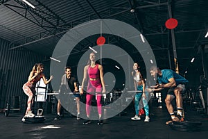 Group of young friends doing sports, training at gym indoors. Sportive woman lifting barbell, bulding muscles