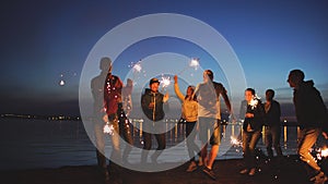 Group of young friends having a beach party. Friends dancing and celebrating with sparklers in twilight sunset