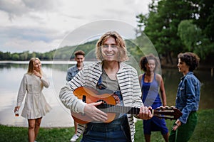 A group of young friends having fun near a lake, laughing and playing guitar.