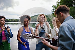 A group of young friends having fun near a lake, laughing and playing guitar.