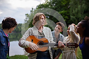 A group of young friends having fun near a lake, laughing and playing guitar.