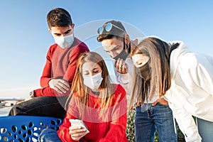 Group of young friends looking at the smartphone outdoor wearing Coronavirus safe protection mask - Two heterosexual couples of