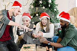 Group of young friends sitting next to a nicely decorated Christmas tree, exchanging Christmas presents and drinkink