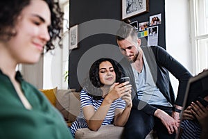 A group of young friends with smartphone sitting on sofa indoors, house sharing concept.