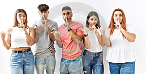 Group of young friends standing together over isolated background asking to be quiet with finger on lips pointing with hand to the