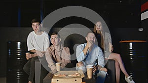 Group of young friends watching football together on couch, communicating, eating popcorn and pizza