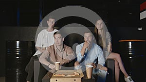 Group of young friends watching television together on couch, communicating, eating popcorn and pizza