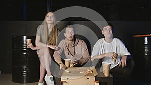 Group of young friends watching television together on couch, communicating, eating popcorn and pizza