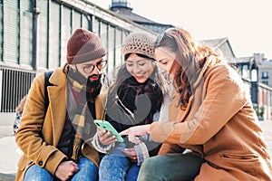 Group of young happy people using a smartphone device outdoors. Three cheerful friends smiling and watching a cellphone