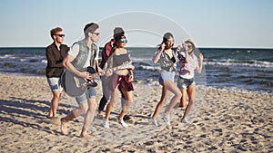 Group of young hipster friends walking and dancing together playing guitar and singing songs on a beach at the water`s