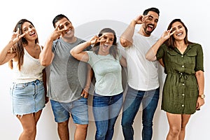 Group of young hispanic friends standing together over isolated background doing peace symbol with fingers over face, smiling